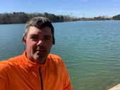 A man in an orange shirt is taking a selfie in front of a lake.