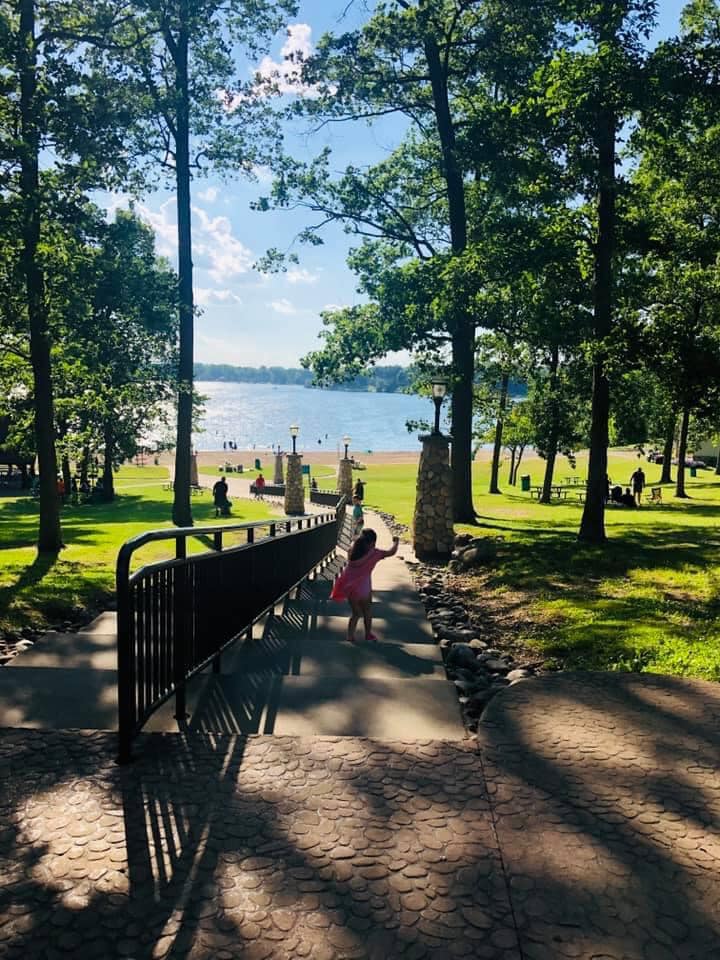 A child in a pink dress runs down stone steps toward a lake, surrounded by trees and a grassy park.