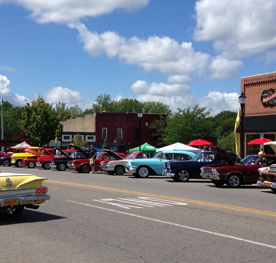 Classic cars parked on a street, likely a car show, sunny day, some buildings and trees in background.