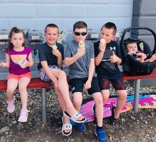 Five children eating ice cream while seated on a bench outside; one baby in a car seat.