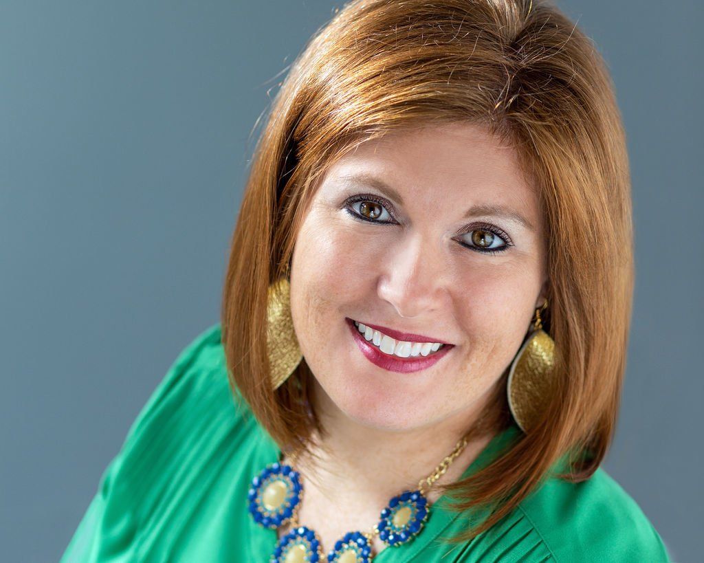 Smiling woman in a green top with blue necklace against a gray background