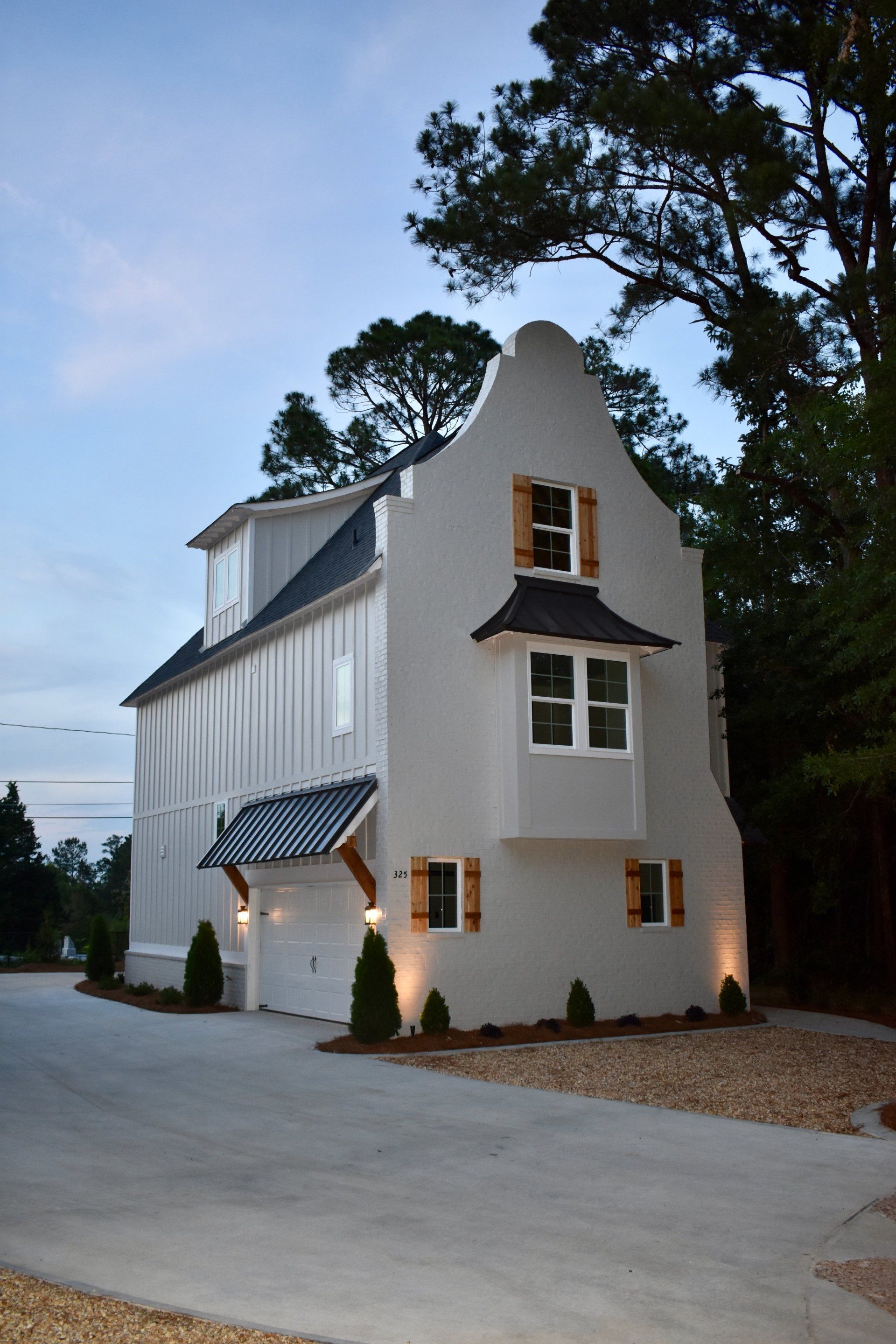 White gabled house with lit windows and blue awning at dusk, beside tall trees and a driveway