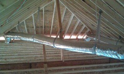 A silver insulated duct runs across exposed wooden rafters in an attic ceiling.