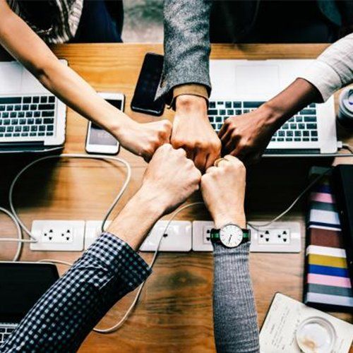 Cinco personas chocando los puños sobre un escritorio con computadoras portátiles, teléfonos y enchufes, promoviendo el trabajo en equipo y la colaboración.