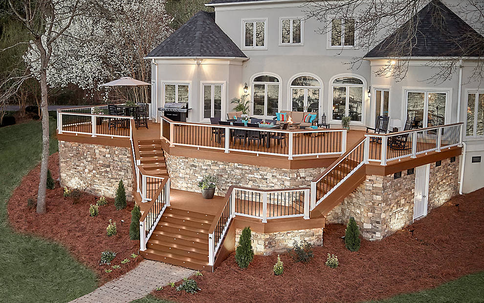 Multi-level deck with stone accents, illuminated, attached to a large house, furnished for outdoor dining and relaxation.