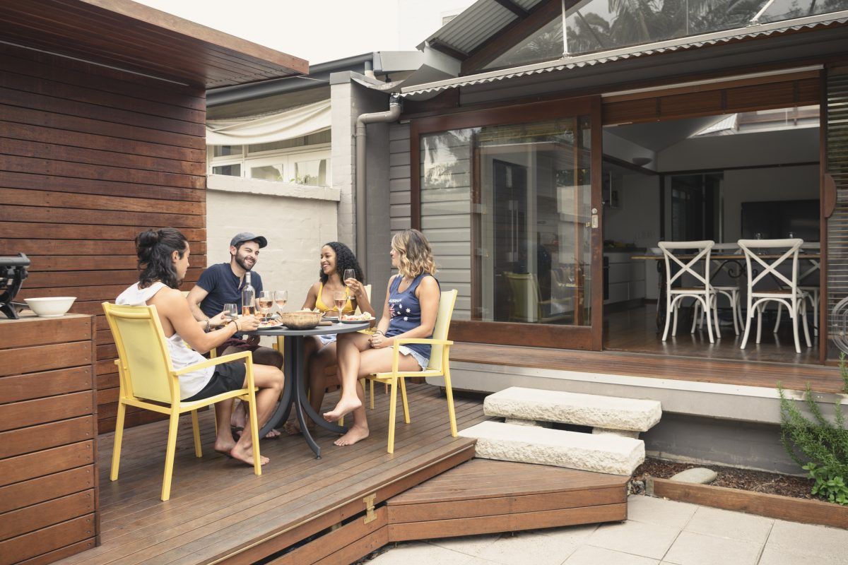 Friends eating at a table on a wooden deck next to a house with open sliding glass doors.