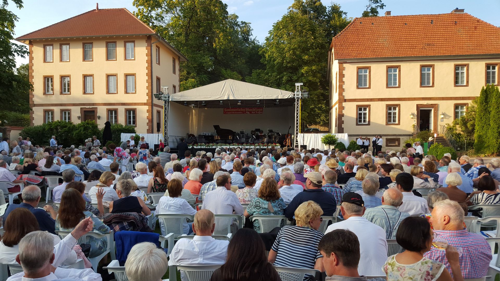 Die Besucher eines Open-Air-Konzerts sitzen an einem sonnigen Tag vor einer Bühne zwischen zwei beigen Gebäuden.
