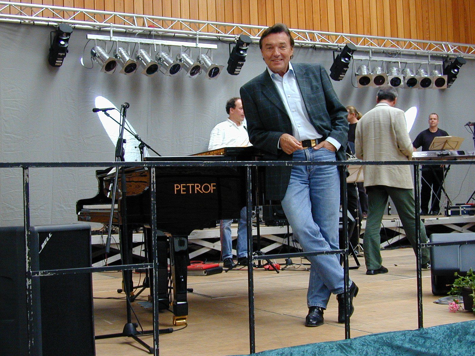 Ein Mann lehnt sich auf einer Bühne an einen Flügel; hinter ihm befinden sich weitere Musiker und Scheinwerfer.