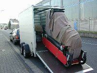 Ein Lieferwagen verlädt einen roten Rollstuhl in einen überdachten Anhänger am Straßenrand.