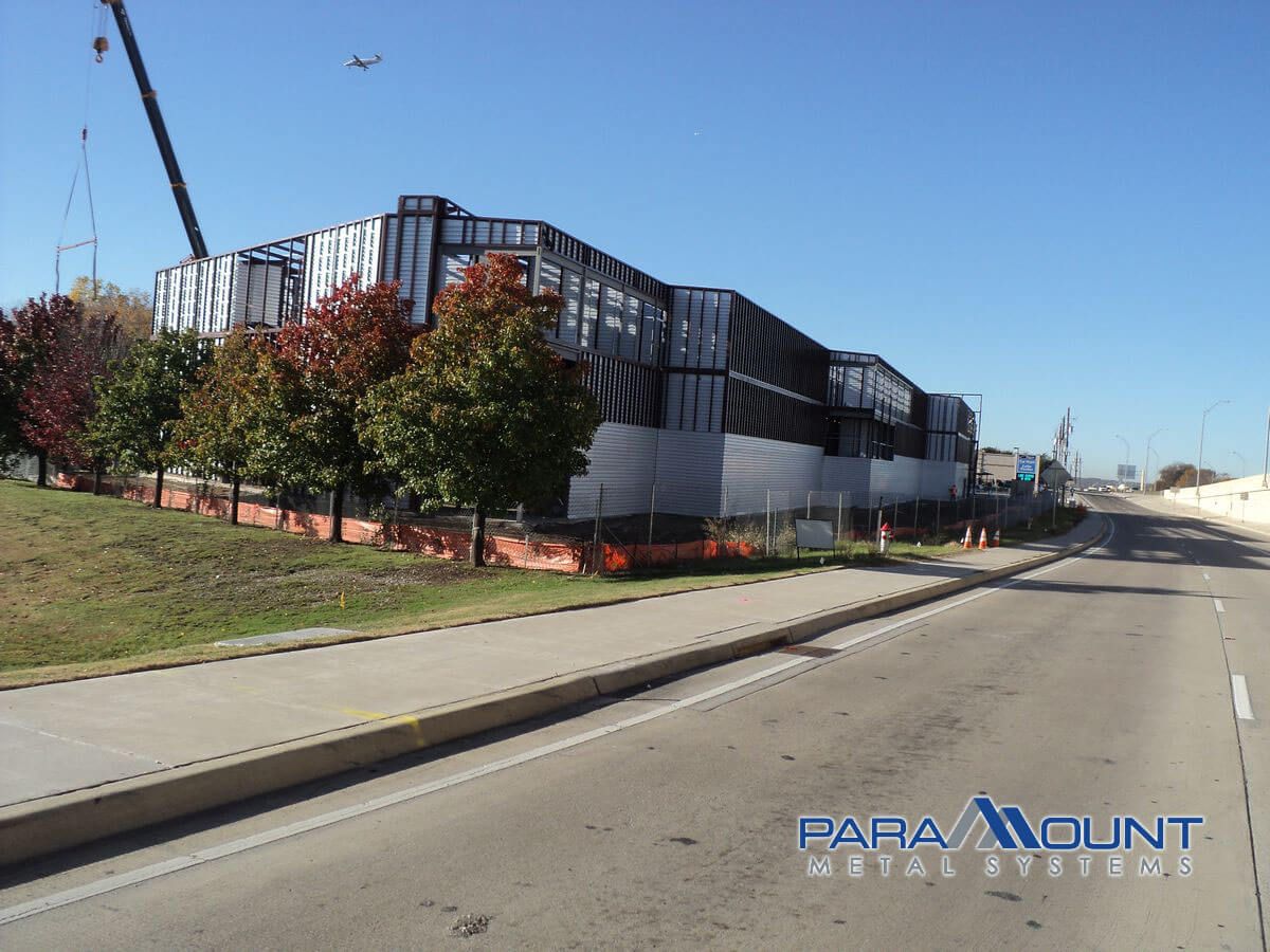 Building under construction; steel frame with silver siding, red brick base, next to road and trees.