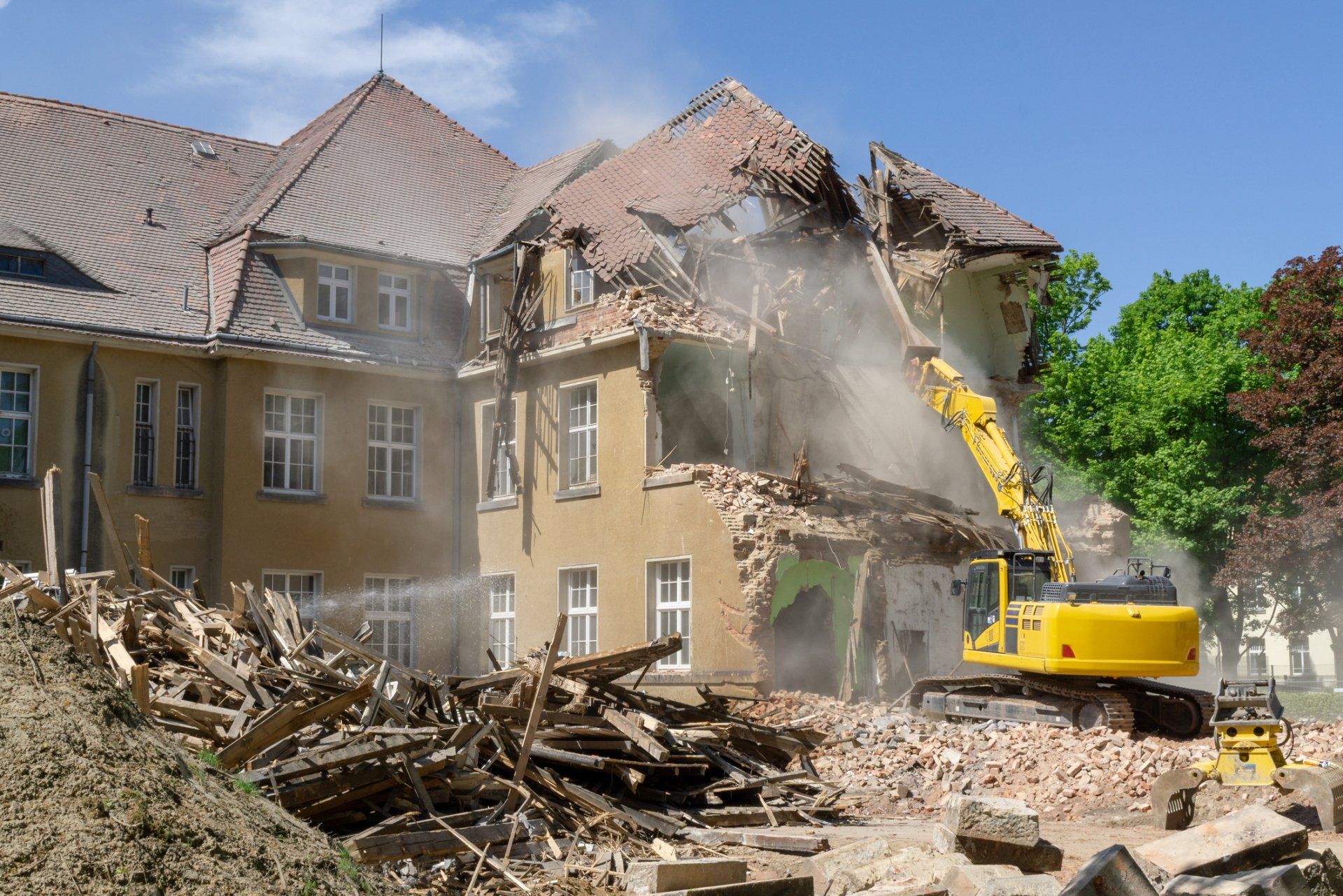 Yellow excavator demolishing a tan building, creating dust and debris; sunny day.