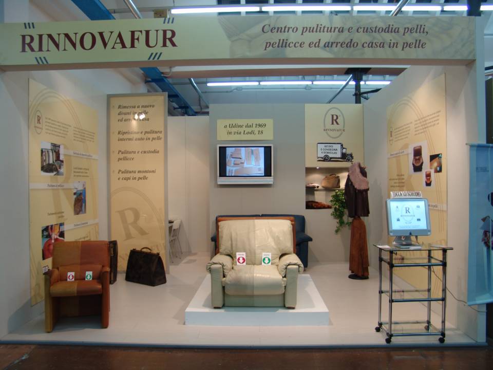 Stand RINNOVAFUR con poltrona beige, vetrine a parete, schermo TV e tavolino in vetro in occasione di una fiera.