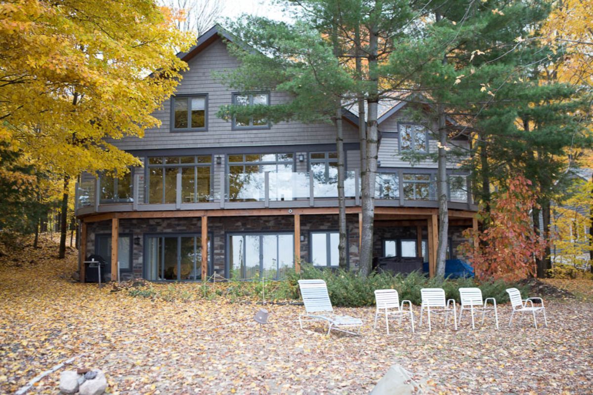 Custom Muskoka Cottage featuring a large house with a lot of windows is surrounded by trees and leaves.