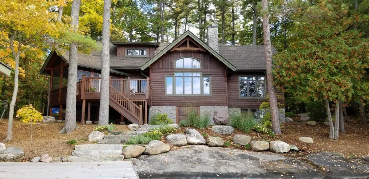 Custom Muskoka Cottage featuring a large log cabin is sitting on the shore of a lake surrounded by trees.