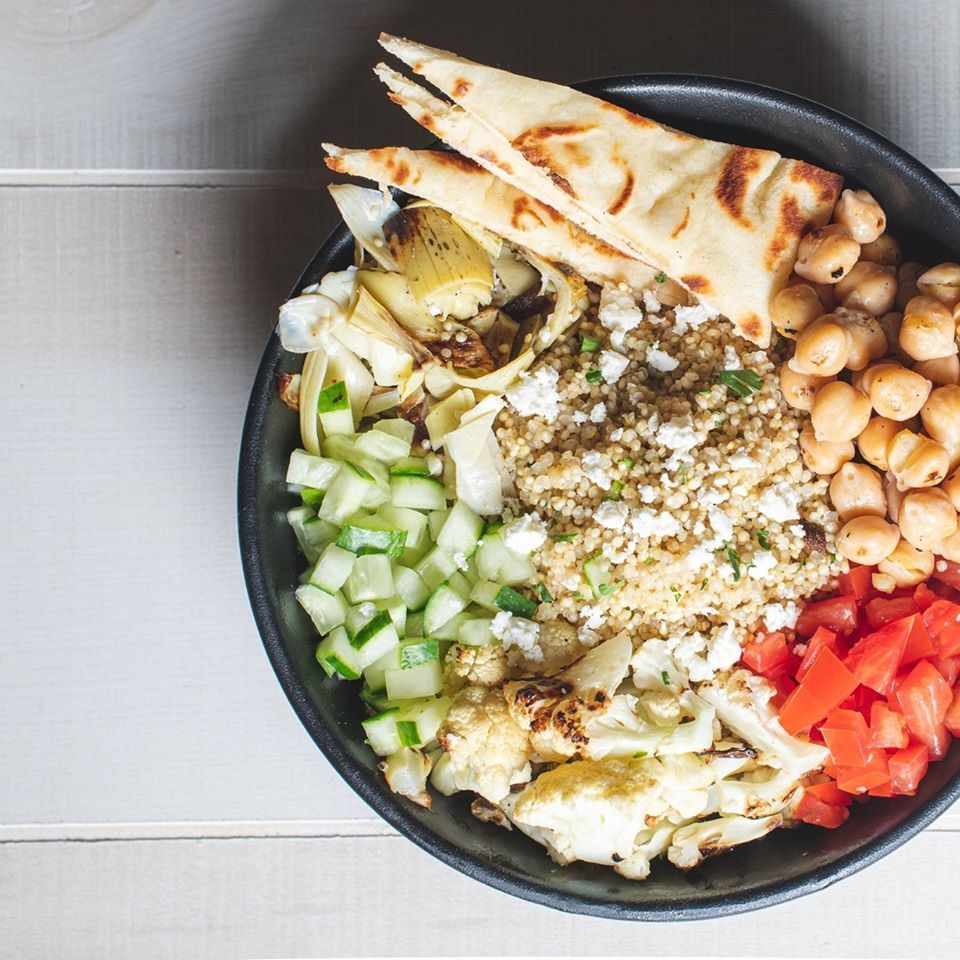 A bowl of food with chickpeas , cucumbers , tomatoes and pita bread on a table.