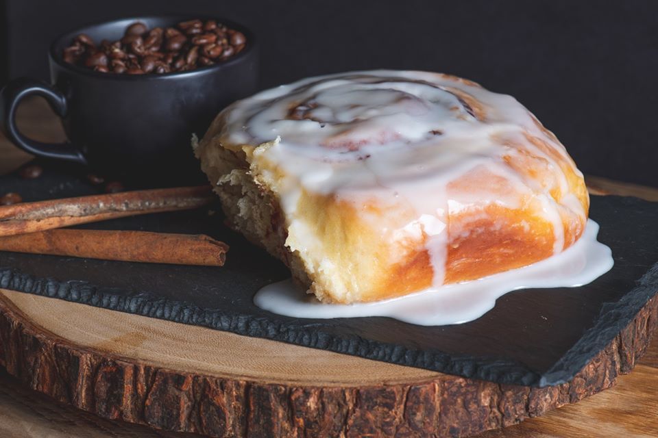 A cinnamon roll with icing on it is on a wooden cutting board next to a cup of coffee.