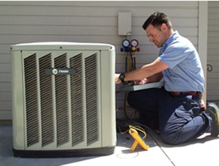 Staff Fixing AC — Kalamazoo, MI — Suburban Heating & Air Conditioning HVAC technician kneels, using tools on an outdoor AC unit. The unit is beige, and the man wears a blue shirt.