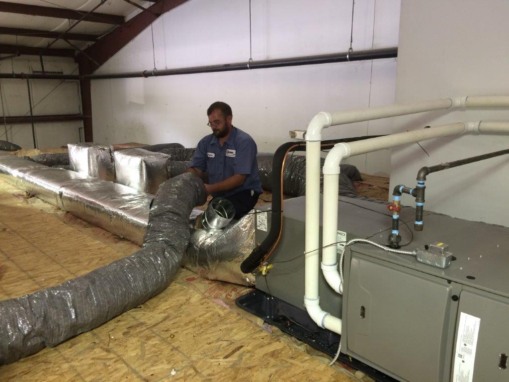 Air Conditioners — Kalamazoo, MI — Suburban Heating & Air Conditioning HVAC technician installing ductwork on a rooftop. Silver ducting, white pipes, grey unit, wood floor.