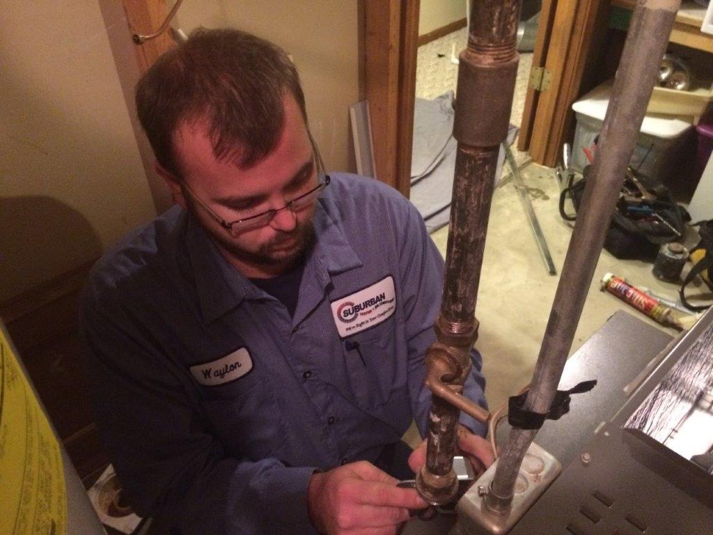 Technician Repairing AC — Kalamazoo, MI — Suburban Heating & Air Conditioning Man in blue work shirt repairs plumbing near furnace.