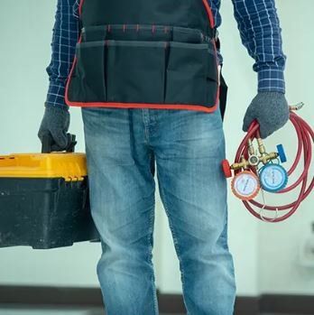 HVAC technician in blue jeans and gloves carries a toolbox and gauges, ready to work.