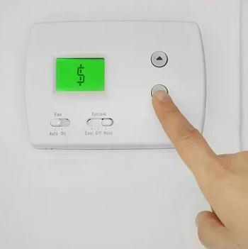 Hand pressing a button on a white thermostat displaying a dollar sign on a green screen.