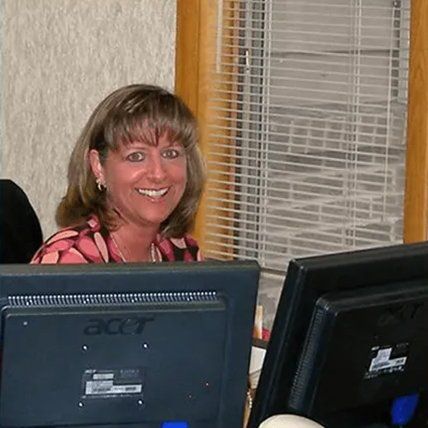 Suburban Staff Smiling — Kalamazoo, MI — Suburban Heating & Air Conditioning Woman smiling at a desk, facing the camera. She's in an office setting with two computer monitors.