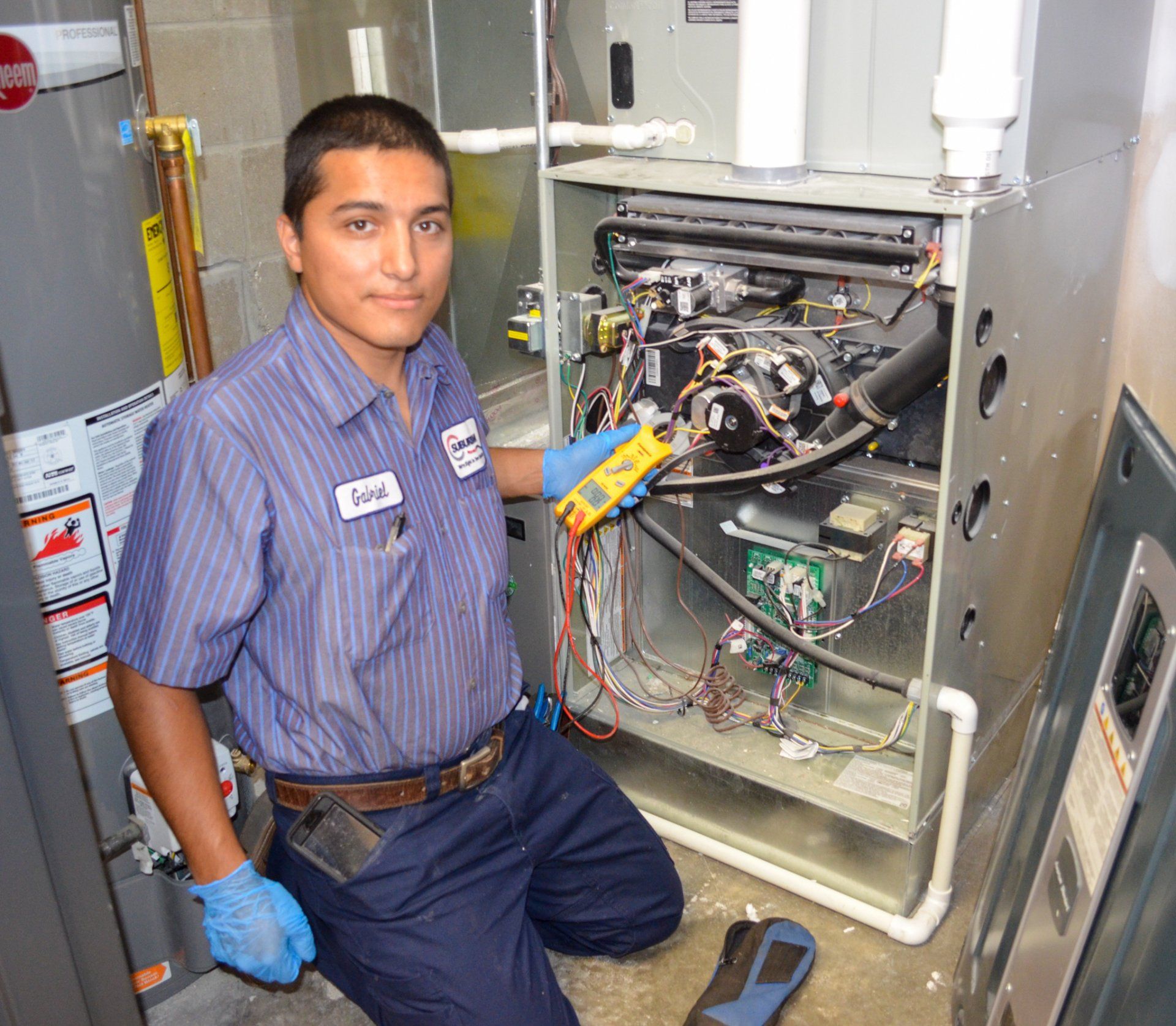 Staff And Customer — Kalamazoo, MI — Suburban Heating & Air Conditioning HVAC technician using multimeter on furnace components. He's wearing a blue uniform and kneeling in a basement.