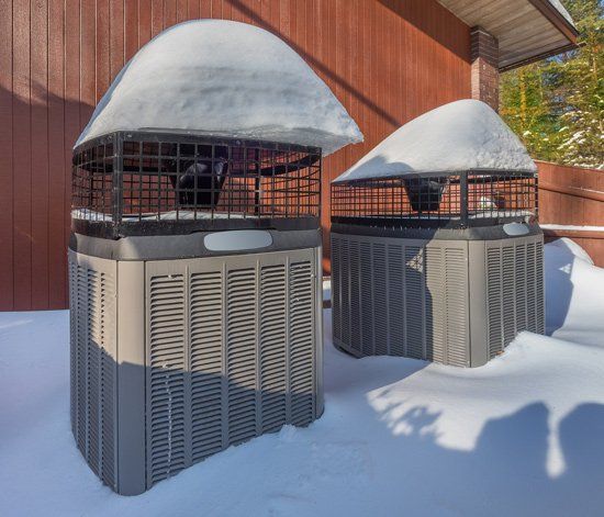 Two Heat Pumps in Winter — Kalamazoo, MI — Suburban Heating & Air Conditioning Two air conditioning units covered with snow in front of a wood-paneled building.