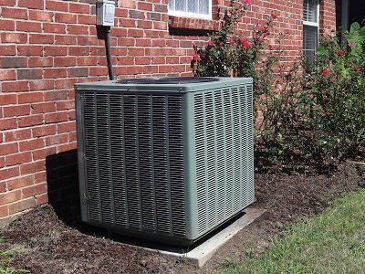 AC Winding Money — Kalamazoo, MI — Suburban Heating & Air Conditioning Air conditioning unit against a brick wall, next to bushes.
