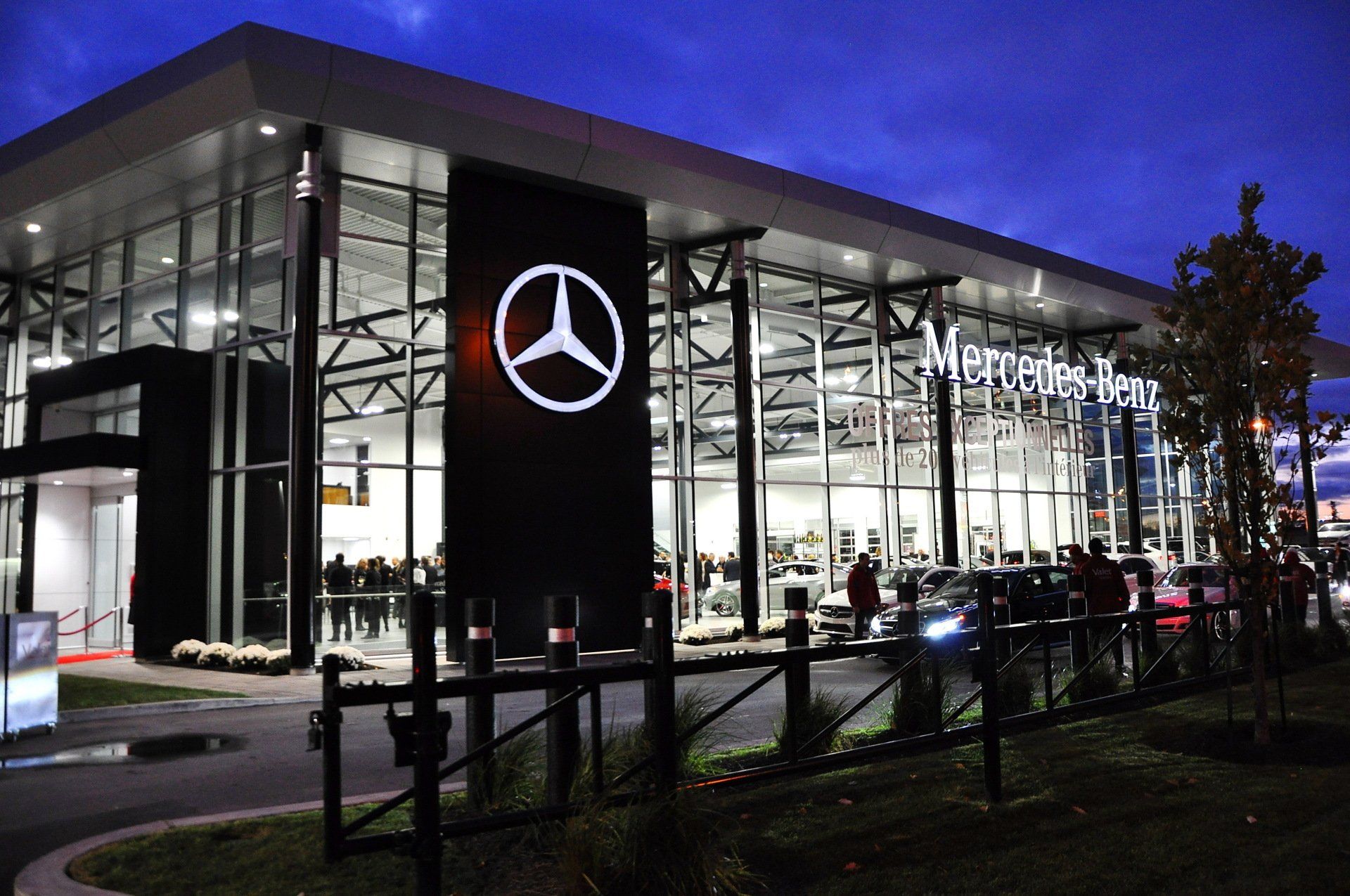 Mercedes-Benz dealership exterior at dusk; illuminated logo and building; cars and people visible.