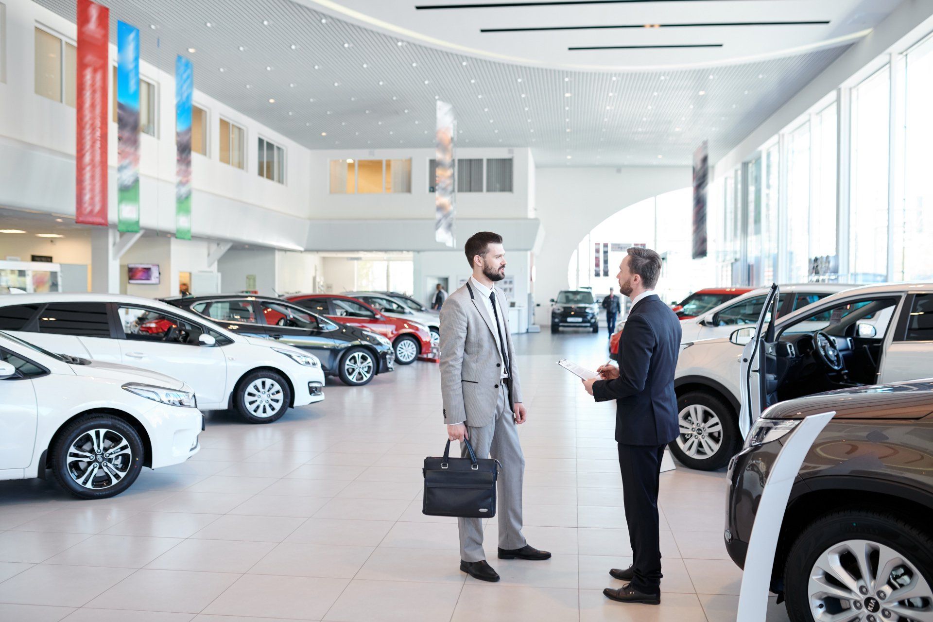 Two men in suits talking in a car dealership, surrounded by various cars.