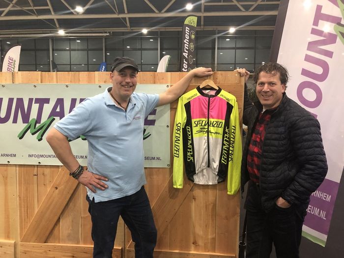 Twee lachende mannen poseren naast een hangend neonkleurig wielershirt bij een stand op een evenement binnen.