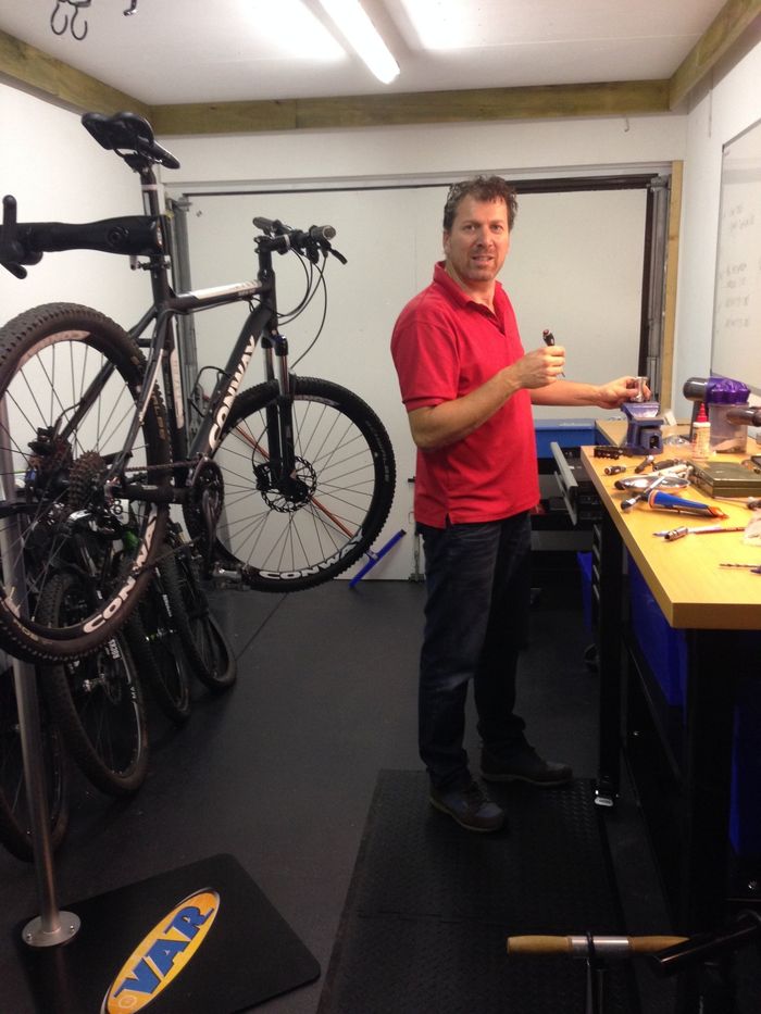 Man in rood shirt met gereedschap in een fietsenwerkplaats, met fietsen aan de muur en onderdelen op een werkbank.