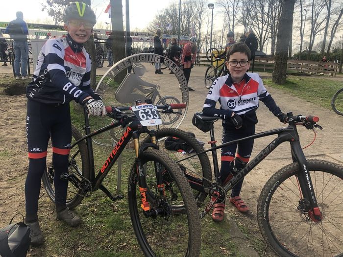 Twee fietsers poseren met mountainbikes op een graspad, gekleed in bijpassende zwarte, witte en rode kleding.
