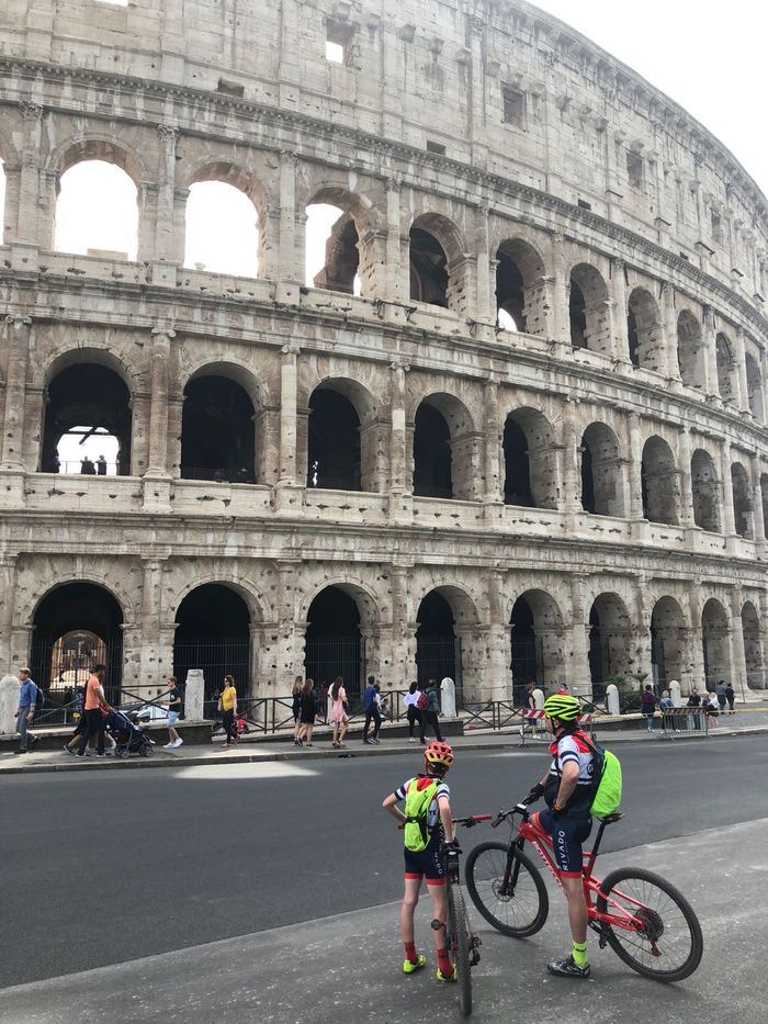 Het Colosseum in Rome met twee fietsers die ervoor stilstaan, waarvan er één een fiets op straat vasthoudt.
