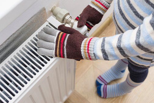 Person in gloves adjusting a white radiator valve. Striped sweater and socks are visible.
