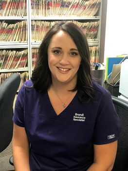 A woman in a purple scrub top is smiling for the camera while sitting at a desk.