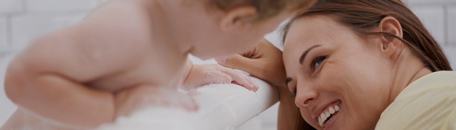 A baby leans toward a smiling person. They appear to be in a light-colored setting, potentially a bathroom.