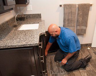 Plumber kneeling by bathroom cabinet, inspecting pipes with a wrench. Dark cabinets, granite countertop, tile floor.
