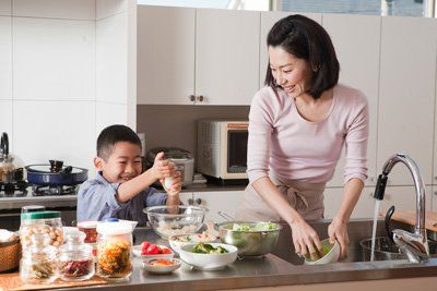 A kitchen that has been repaired by a kitchen plumber in Jackson, MI Woman and child in kitchen, preparing food; woman washing dishes, child mixing ingredients, smiling.