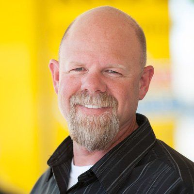 plumbing guarantee Bald man with a trimmed, gray beard, smiles and wears a black collared shirt in front of a yellow background.