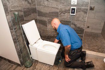 Plumber kneels, repairing a white toilet in a bathroom with gray tile walls.