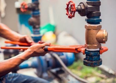 Hands using a wrench to tighten a pipe fitting with a red valve in an outdoor setting.