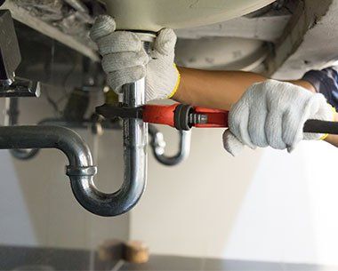 Plumber Fixing White Sink Pipe — Holt, MI — Michigan Plumbing Hands of a plumber in gloves, using a wrench on a sink drainpipe.