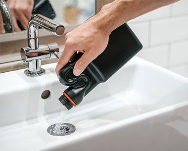 Worker Cleaning Drain — Grand Ledge — Michigan Plumbing Hand pouring liquid from a black bottle into a white sink drain in a bathroom.