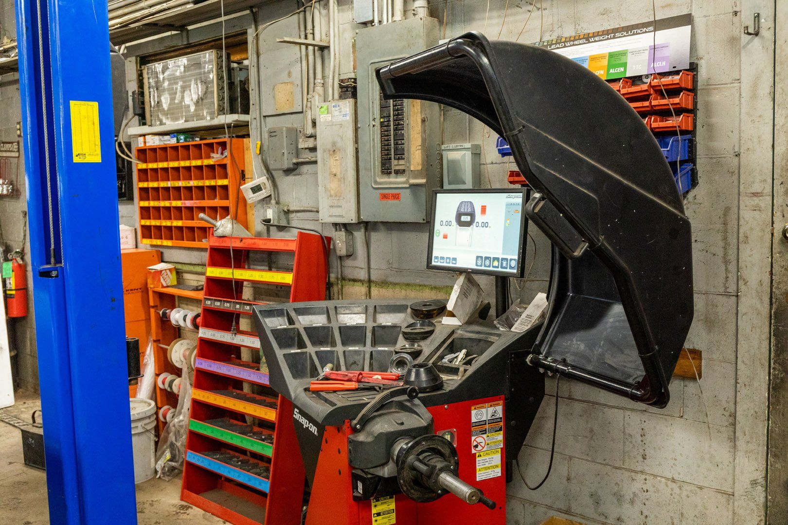 Wheel balancing machine in a garage, with monitor showing wheel data. Shelves with parts visible.