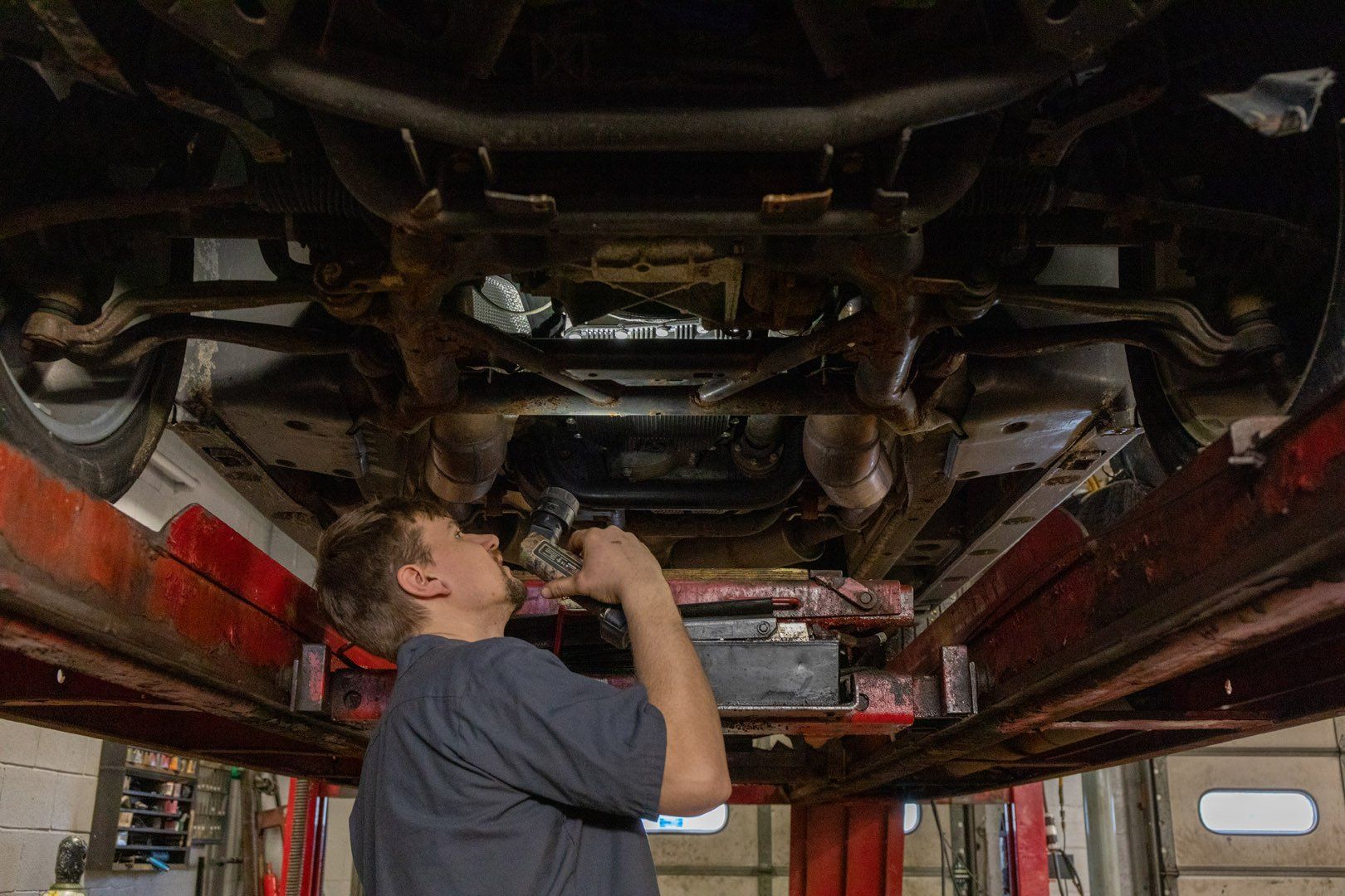 Mechanic inspecting underside of car on a lift. He's holding a light, looking up, in a garage.