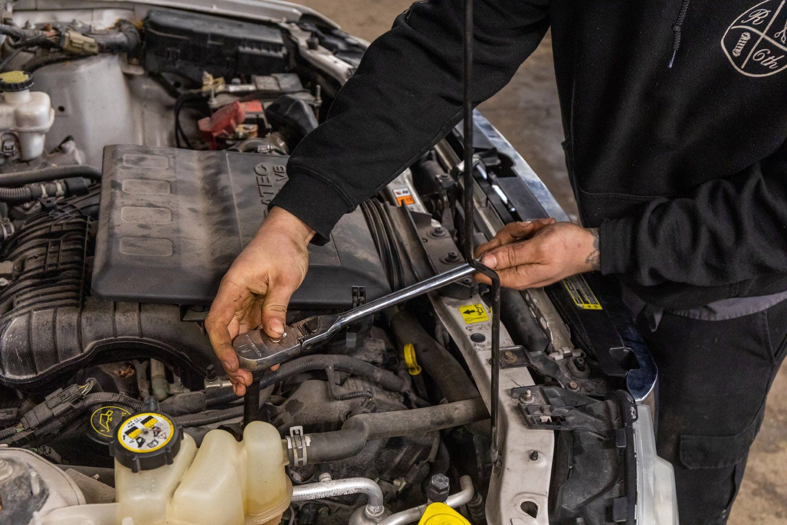 Person in black hoodie working on a car engine, using a tool.