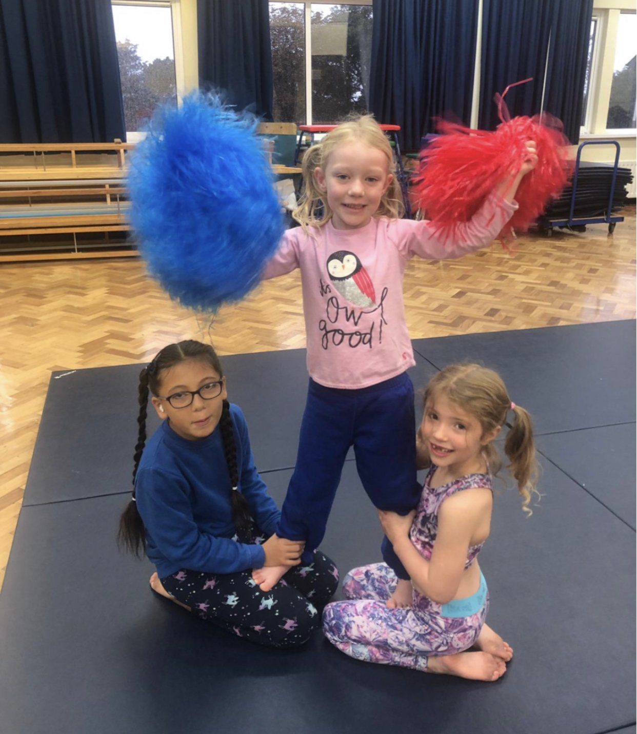 Three little girls are sitting on a mat holding pom poms