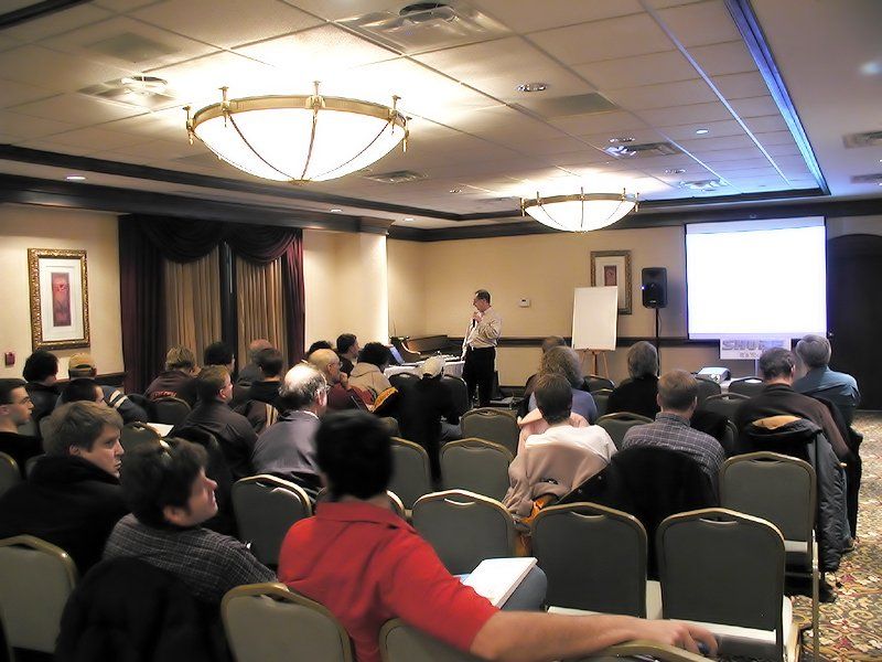 People in chairs facing a presenter at a conference. The room has beige walls, a screen, and overhead lights.